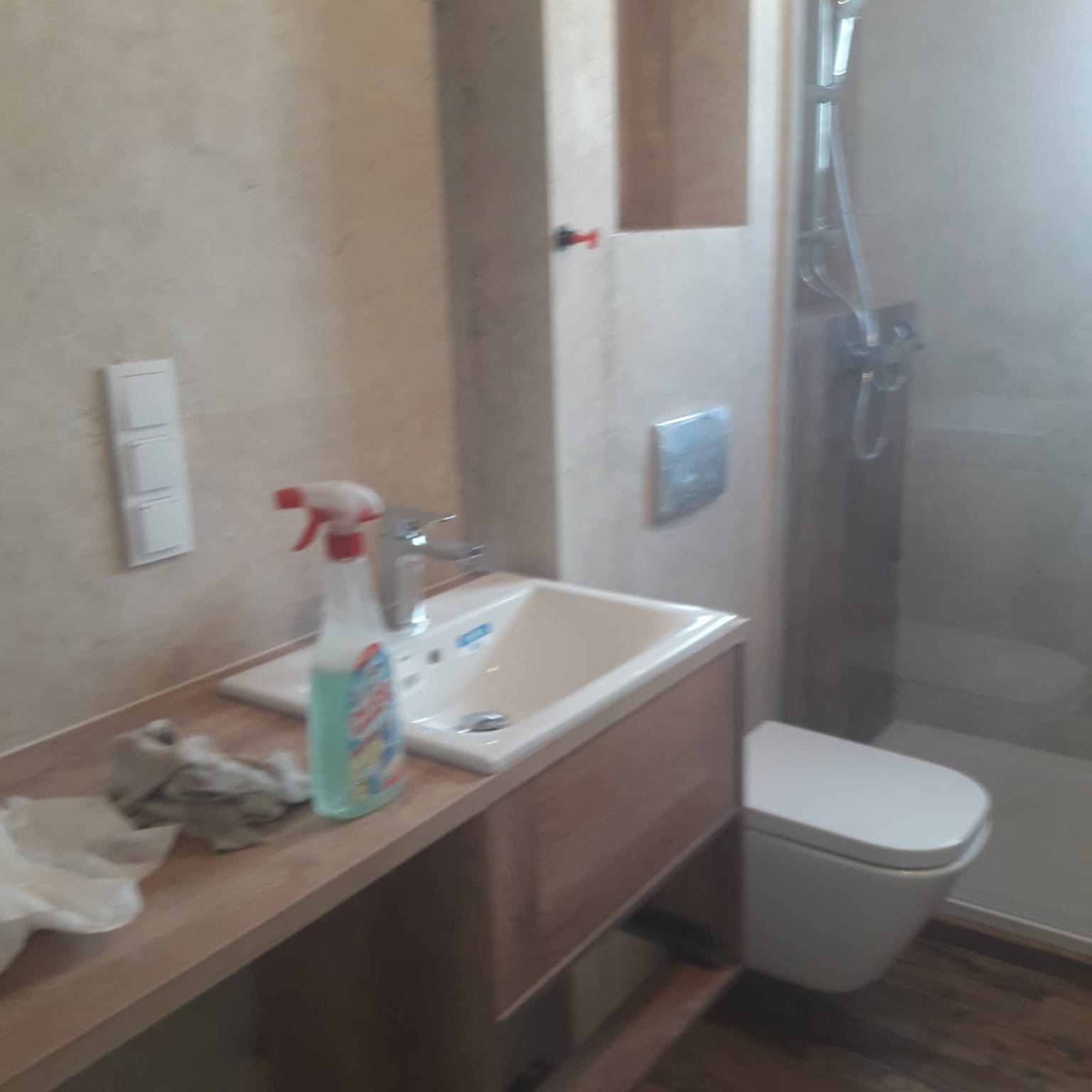 Umywalka z szafką w jasnej łazience z widoczną kabiną prysznicową, na blacie leży szmatka i stoi butelka środka czyszczącego.