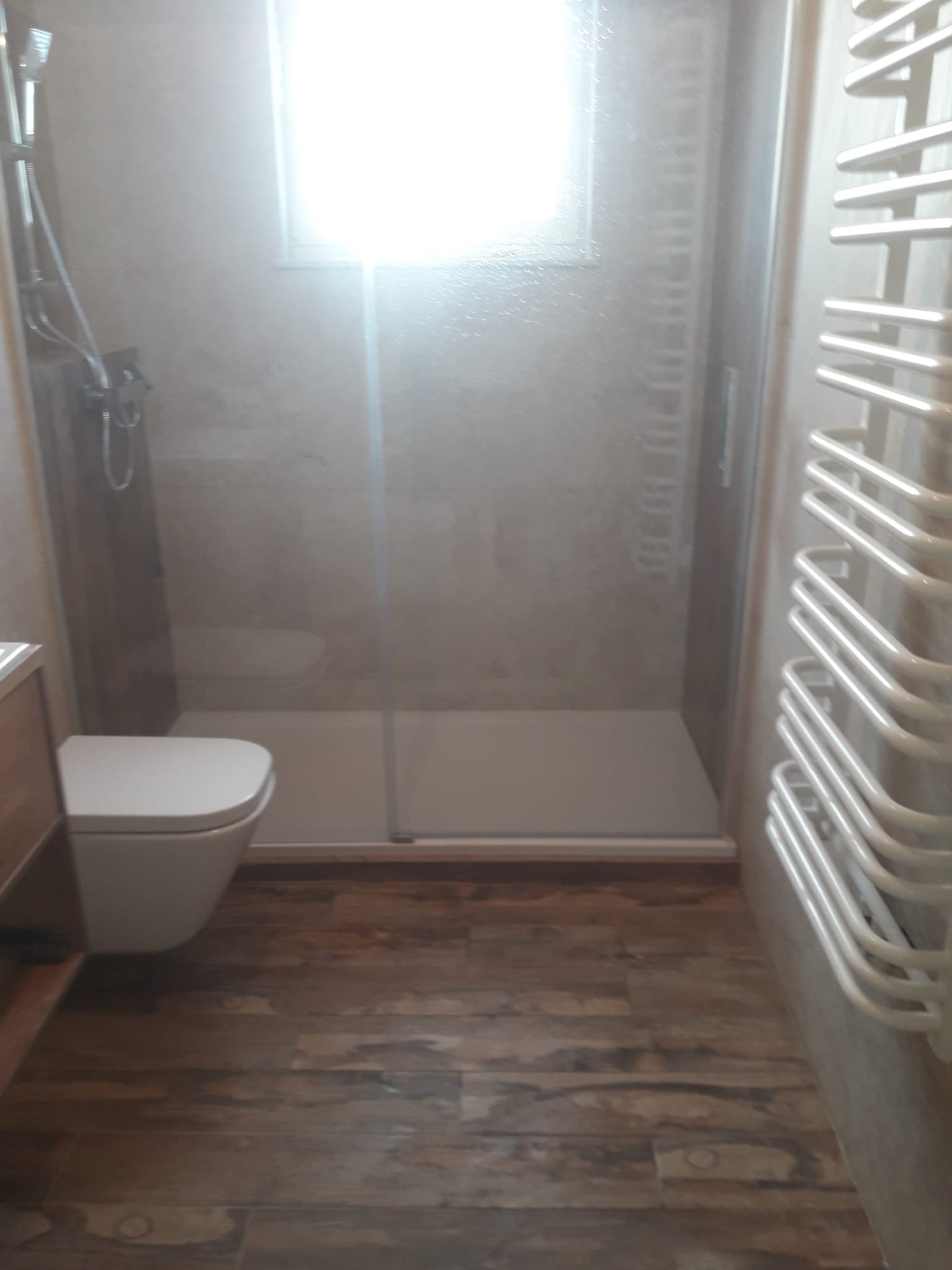 Wykończona łazienka z kabiną prysznicową typu walk-in, wiszącą toaletą i grzejnikiem drabinkowym, wyłożona płytkami imitującymi drewno na podłodze i beżowymi płytkami na ścianach.