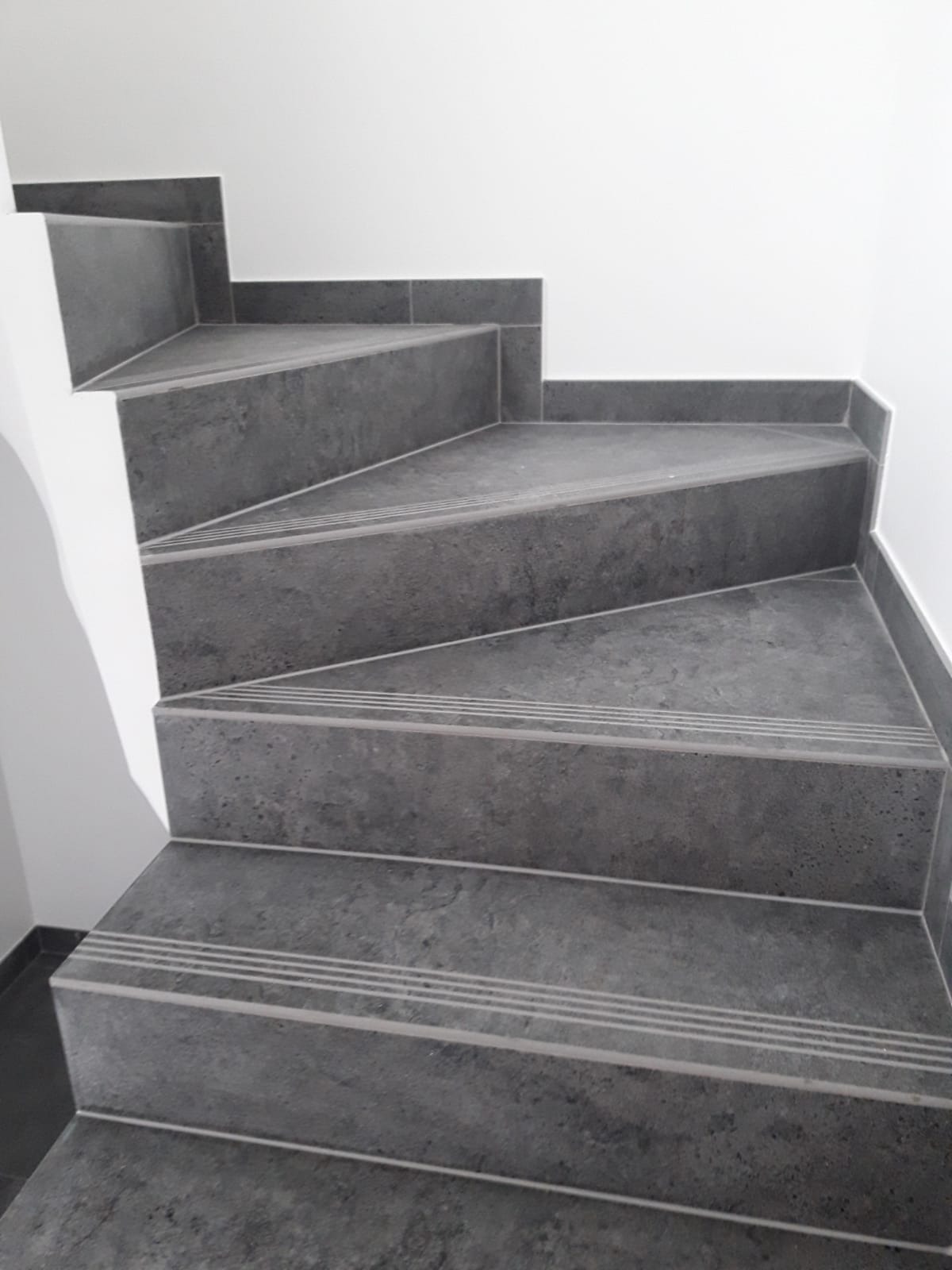 Schody wyłożone szarą, porowatą płytką z trzema antypoślizgowymi paskami na krawędzi stopnia, widoczne białe ściany po bokach.