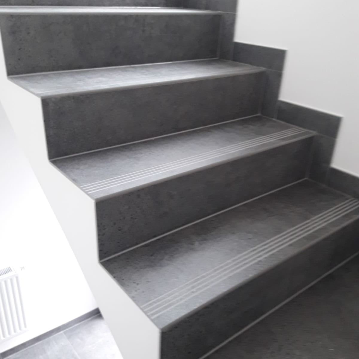 Schody wyłożone ciemnoszarą płytką z antypoślizgowymi paskami, widoczne fragmenty białej ściany i grzejnika.