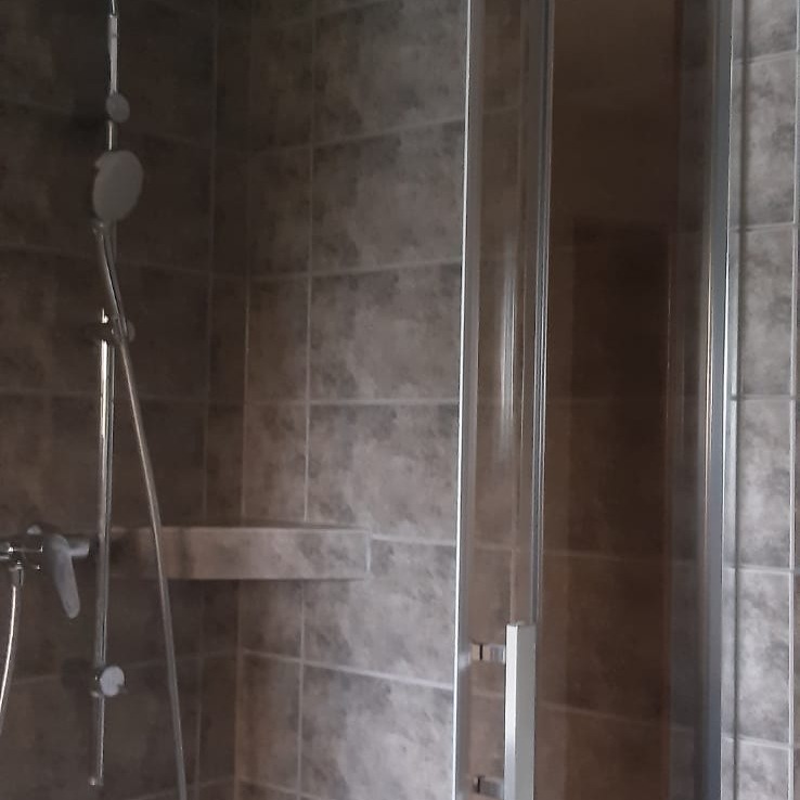 Kabina prysznicowa z przesuwnymi drzwiami, wykończona szarą, kwadratową glazurą; widoczna półka wkomponowana w płytki i chromowana bateria z deszczownicą.