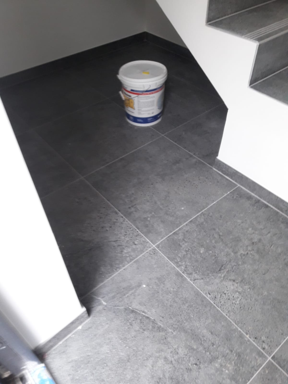 Ciemnoszare płytki podłogowe ułożone pod schodami, z widocznym wiaderkiem z farbą lub klejem, sugerujące prace wykończeniowe.