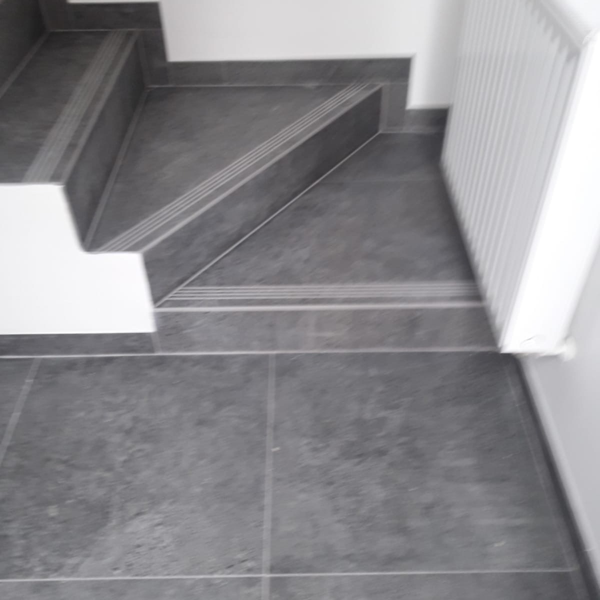 Szare płytki na schodach z antypoślizgowymi paskami i grzejnikiem na ścianie, widok z góry.
