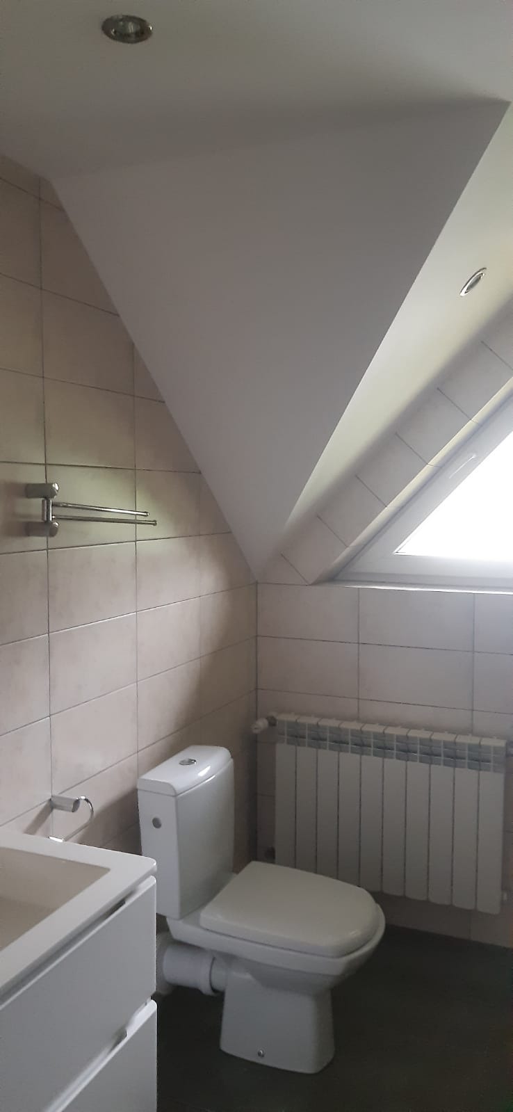 Ukończona łazienka na poddaszu ze skośnym sufitem, beżowe płytki, biała armatura i grzejnik pod oknem.