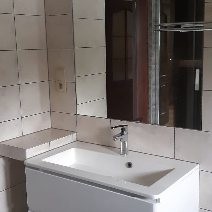 Umywalka nablatowa z białą szafką, chromowaną baterią i lustrem w ramie na tle beżowych, kwadratowych płytek. Widoczna półka wyłożona tymi samymi płytkami.