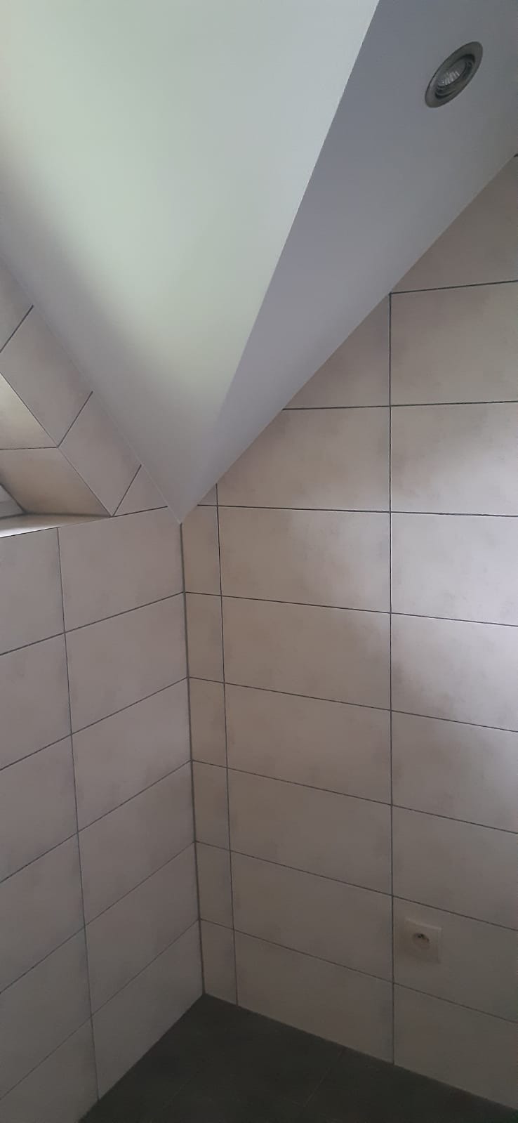 Fragment wykończonej łazienki z beżowymi, prostokątnymi płytkami na ścianach, ciemną podłogą i skośnym sufitem z wbudowanym oświetleniem punktowym.