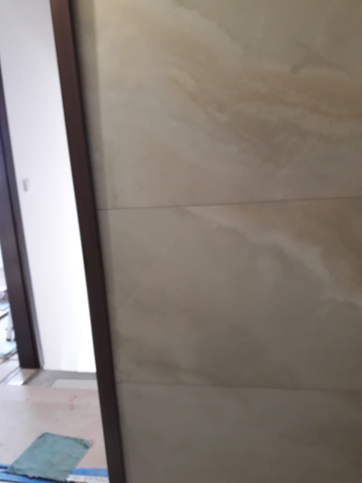 Beżowe płytki imitujące marmur na ścianie, widoczne łączenia, fragment futryny drzwi i podłogi z niebieską szmatką w tle.