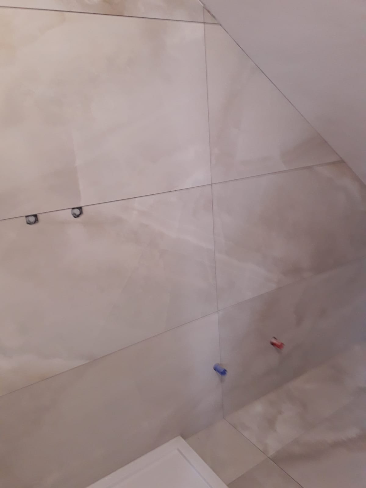 Ściana wyłożona jasnymi, marmurkowymi płytkami. Widoczne krzyżyki dystansowe w spoinach, czerwony i niebieski. Ujęcie z dołu, ukazujące fragment sufitu i obudowy wanny.