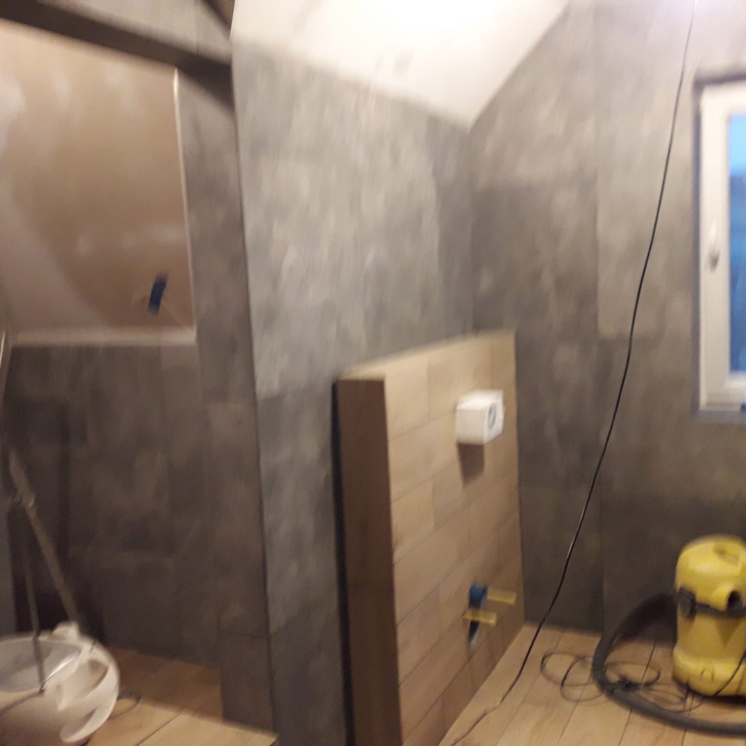 Łazienka w trakcie remontu: szare płytki na ścianach, drewnopodobne płytki na podłodze, zamontowana podtynkowa spłuczka WC, widoczny odkurzacz przemysłowy i wiadro z narzędziami.