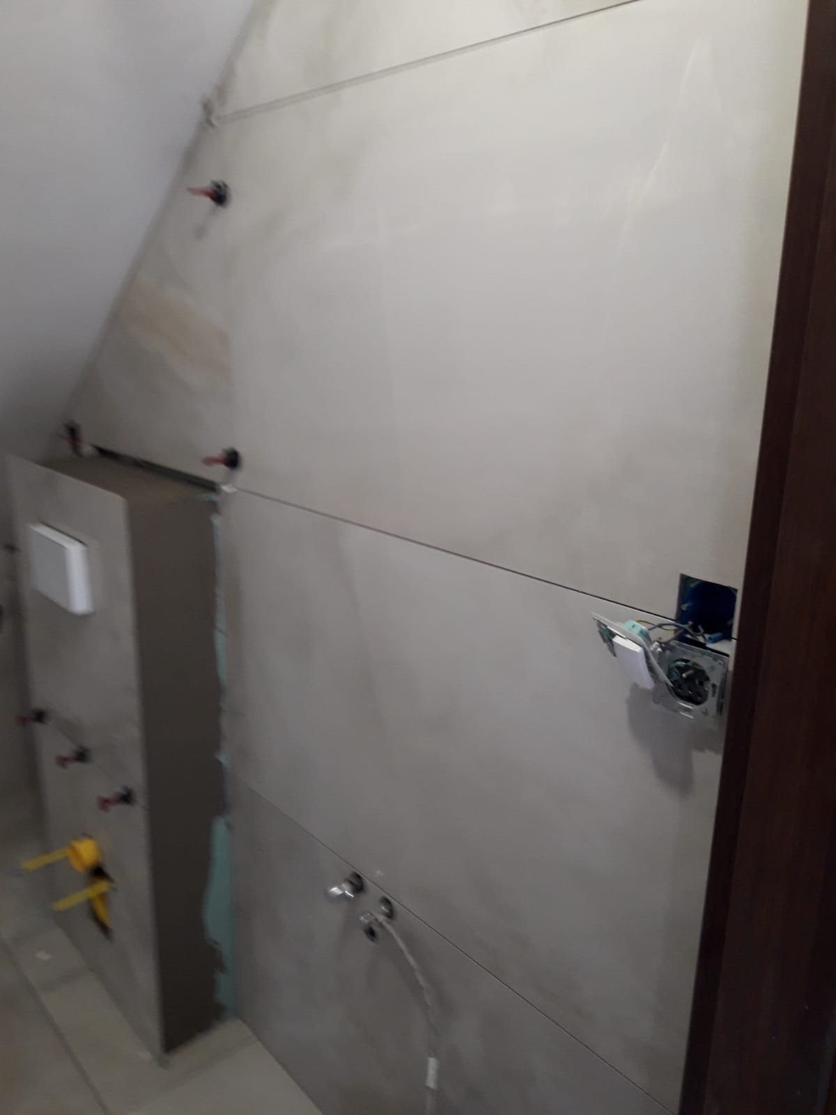 Montaż szarych, matowych płytek w łazience na poddaszu, widoczne otwory na armaturę, puszka elektryczna z wystającymi przewodami, oraz system podtynkowy WC w trakcie instalacji.