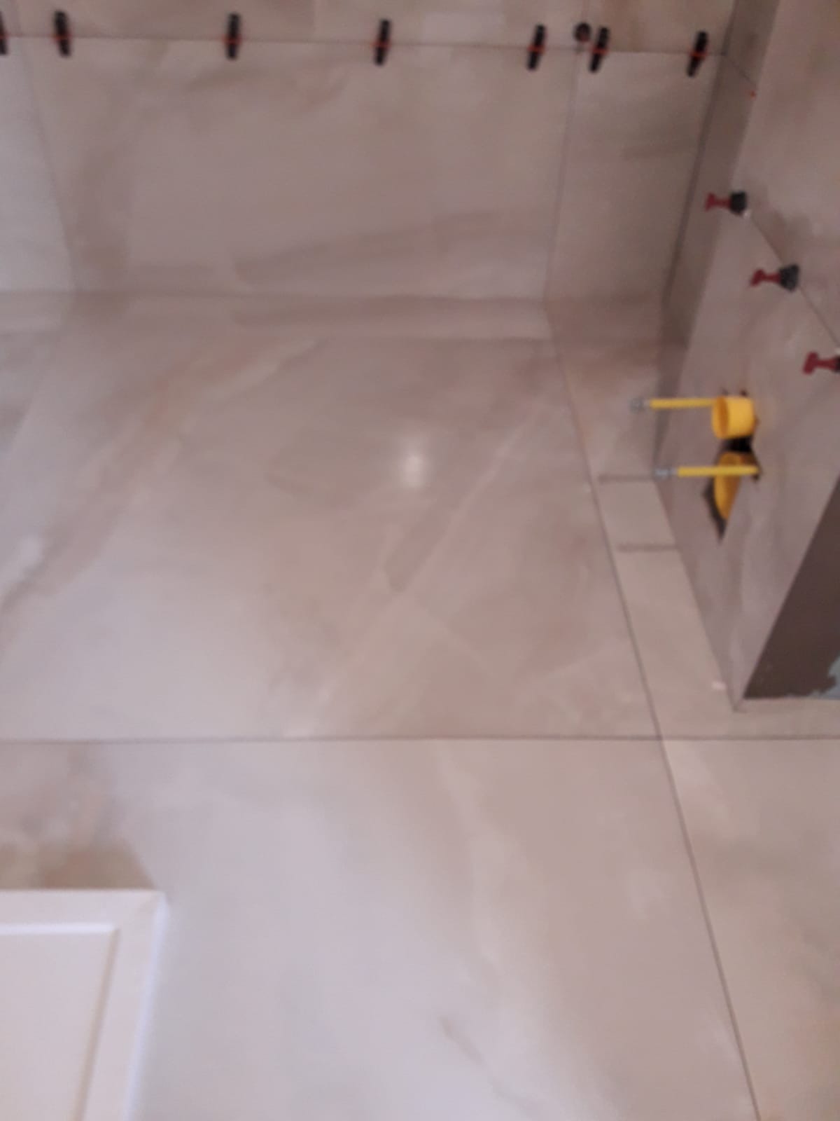 Świeżo ułożone, beżowe płytki podłogowe i ścienne z widocznymi systemami poziomowania płytek na ścianie, w trakcie prac wykończeniowych w łazience.