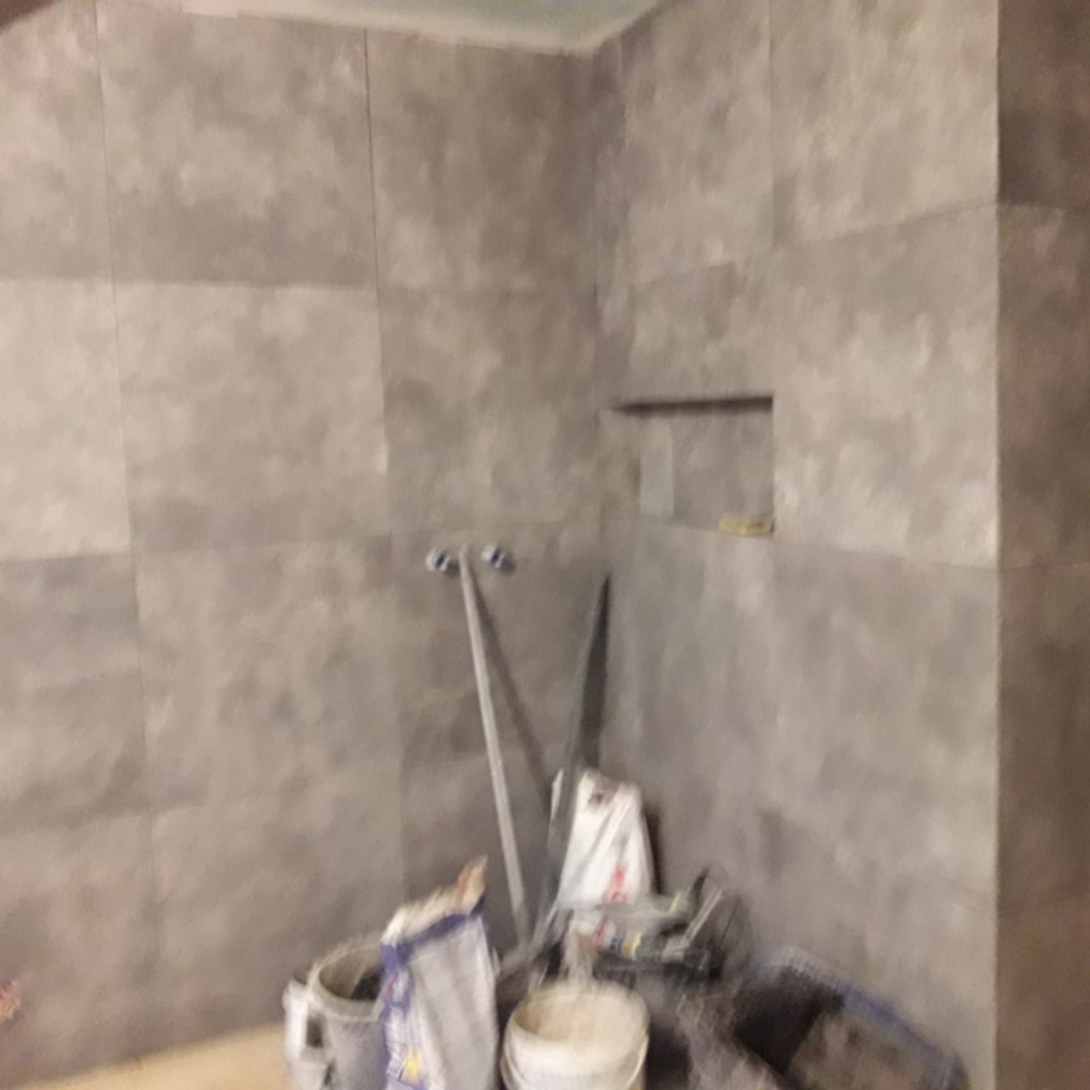 Ściana łazienki wyłożona szarymi płytkami w trakcie prac, widoczne narzędzia i materiały budowlane, półka w ścianie.