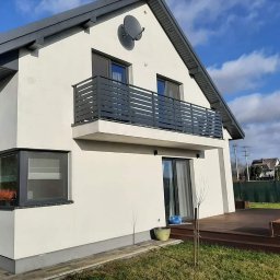 KAM-Weld KAMIL DOROSZ - Nowoczesny dom z balkonem z szarą metalową balustradą poziomą, widok z ogrodu, drewniany taras i zadbany trawnik.