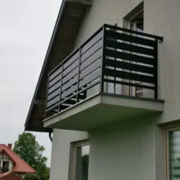 Nowoczesny balkon z czarną, poziomą balustradą z metalowych profili, widok z dołu, w tle dom z czerwoną dachówką.