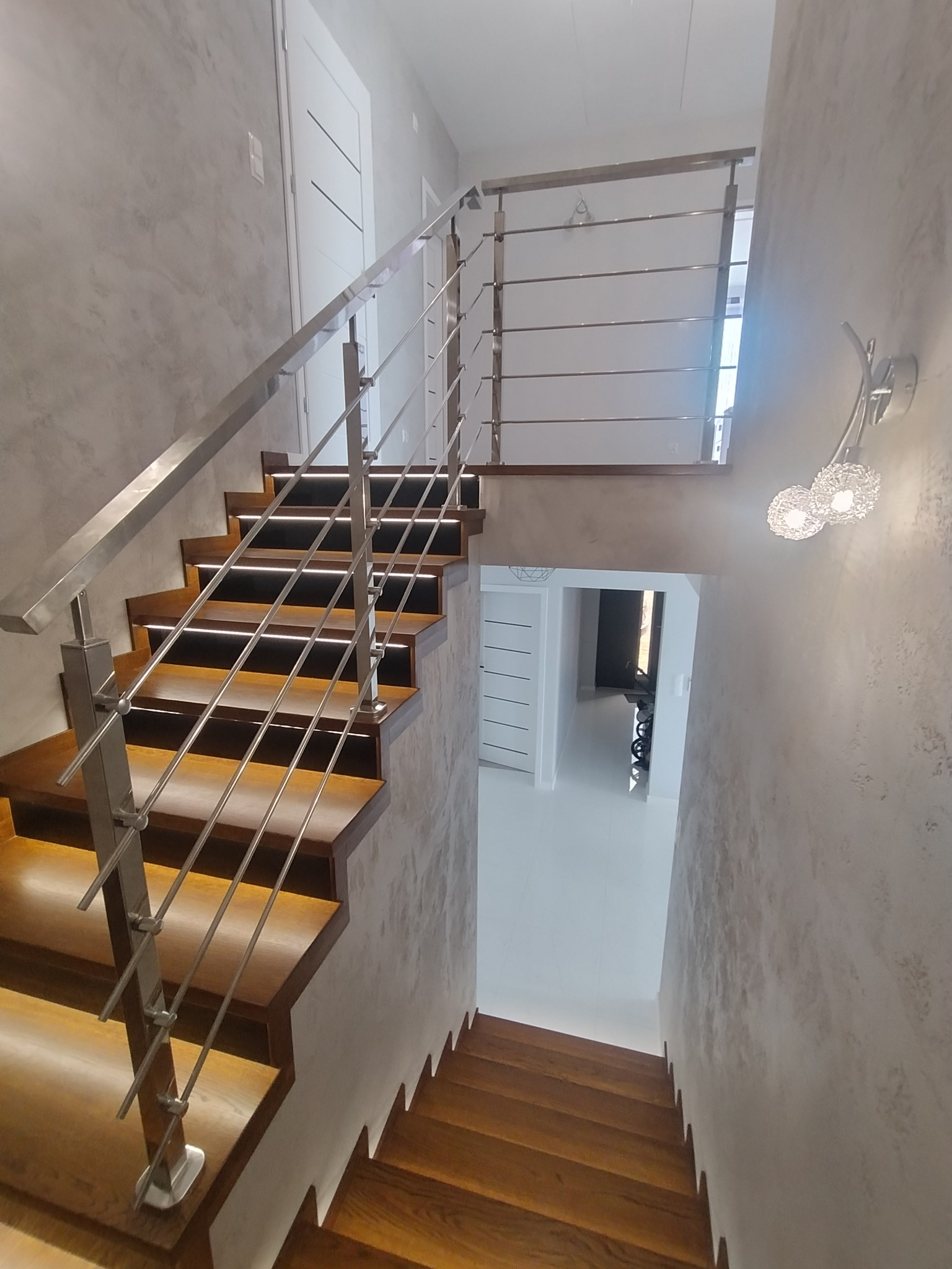 Nowoczesne schody z drewnianymi stopniami podświetlonymi LED i balustradą ze stali nierdzewnej, widok z góry, ściany wykończone w stylu betonu architektonicznego, w tle widoczne białe drzwi.