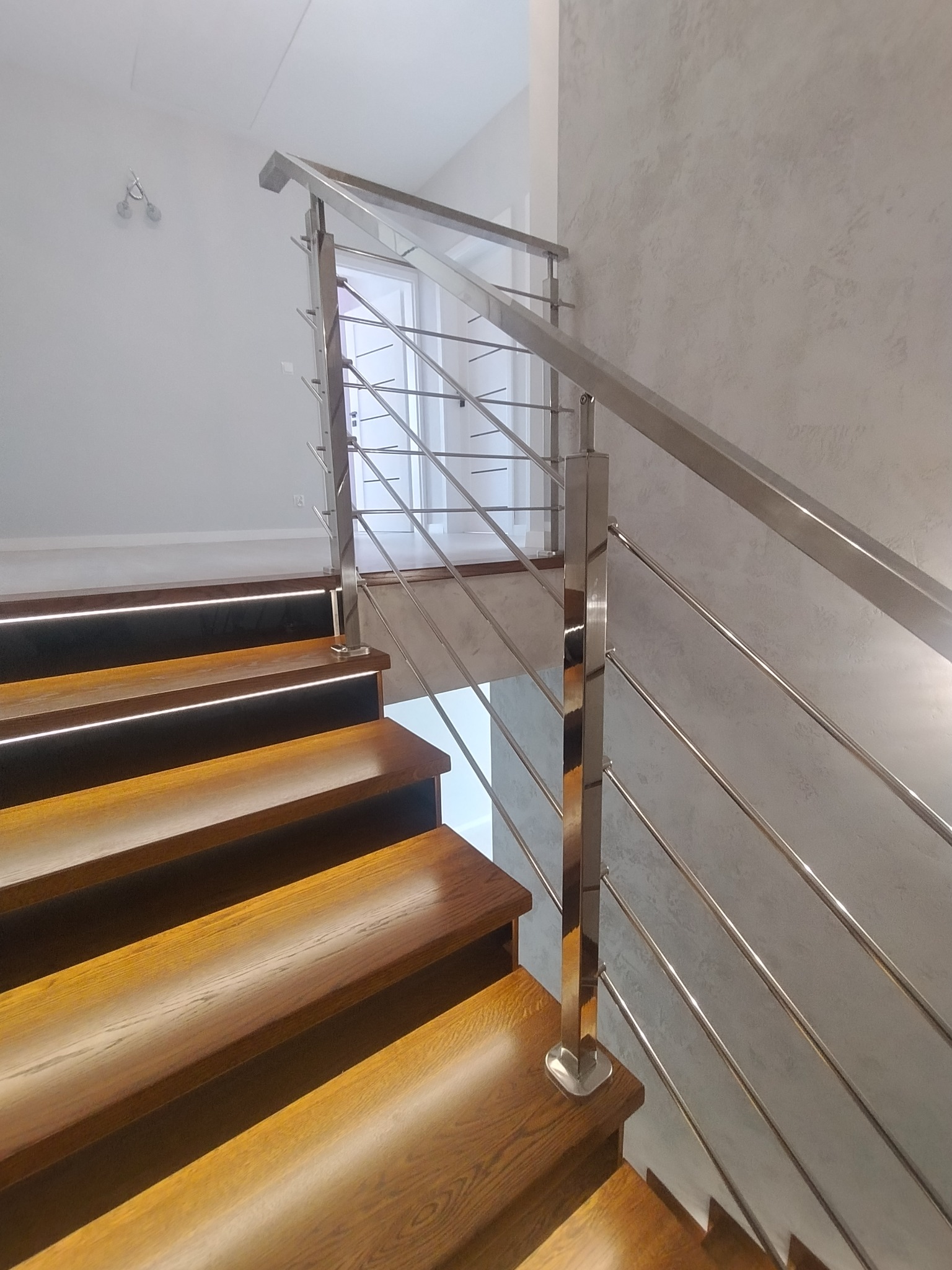 Drewniane schody z podświetleniem LED na stopniach i nowoczesną balustradą ze stali nierdzewnej, widok z dołu, ściana o fakturze betonu.