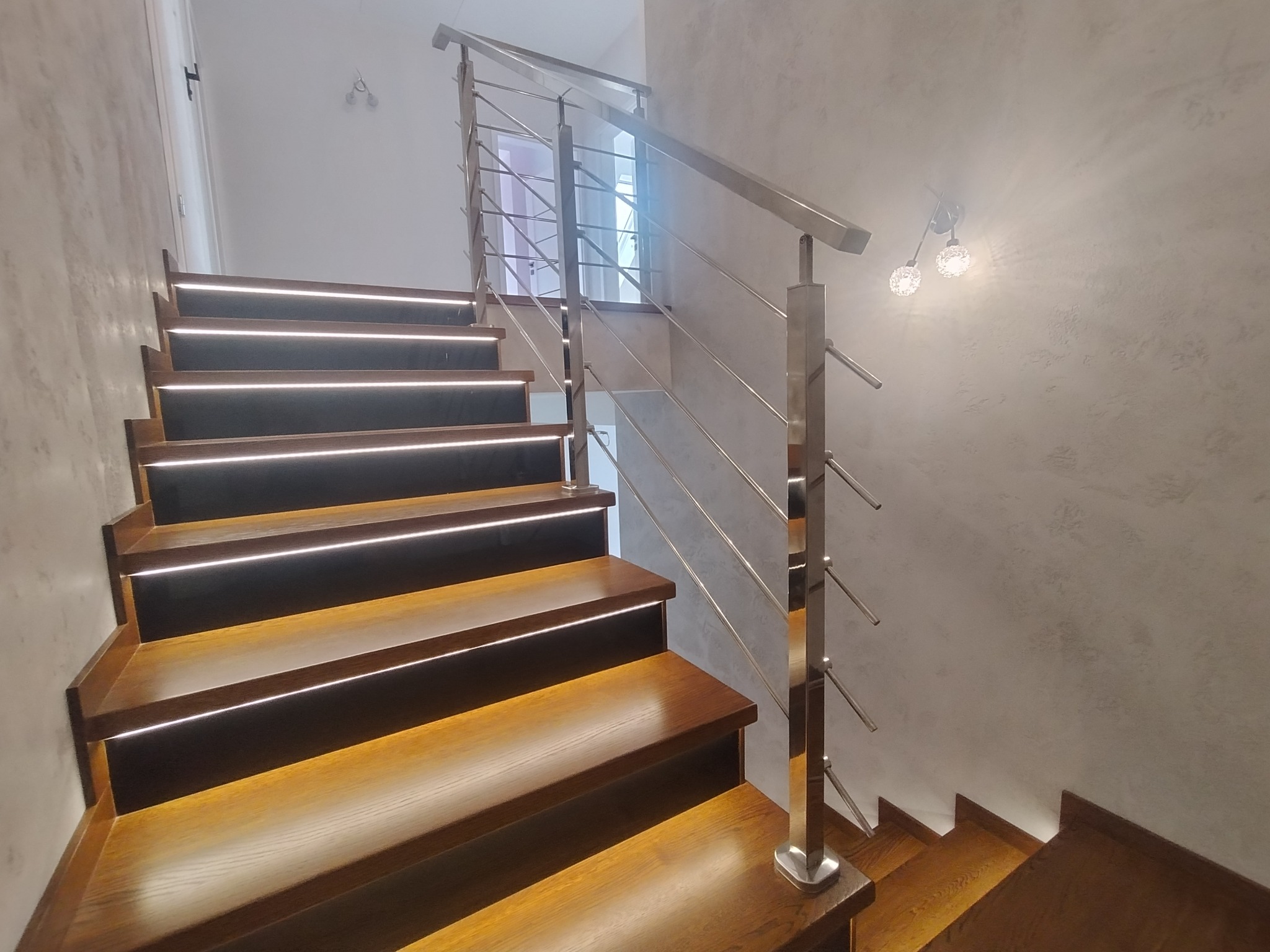 Drewniane schody z podświetleniem LED i balustradą ze stali nierdzewnej, widok z dołu, na ścianie dekoracyjny tynk.