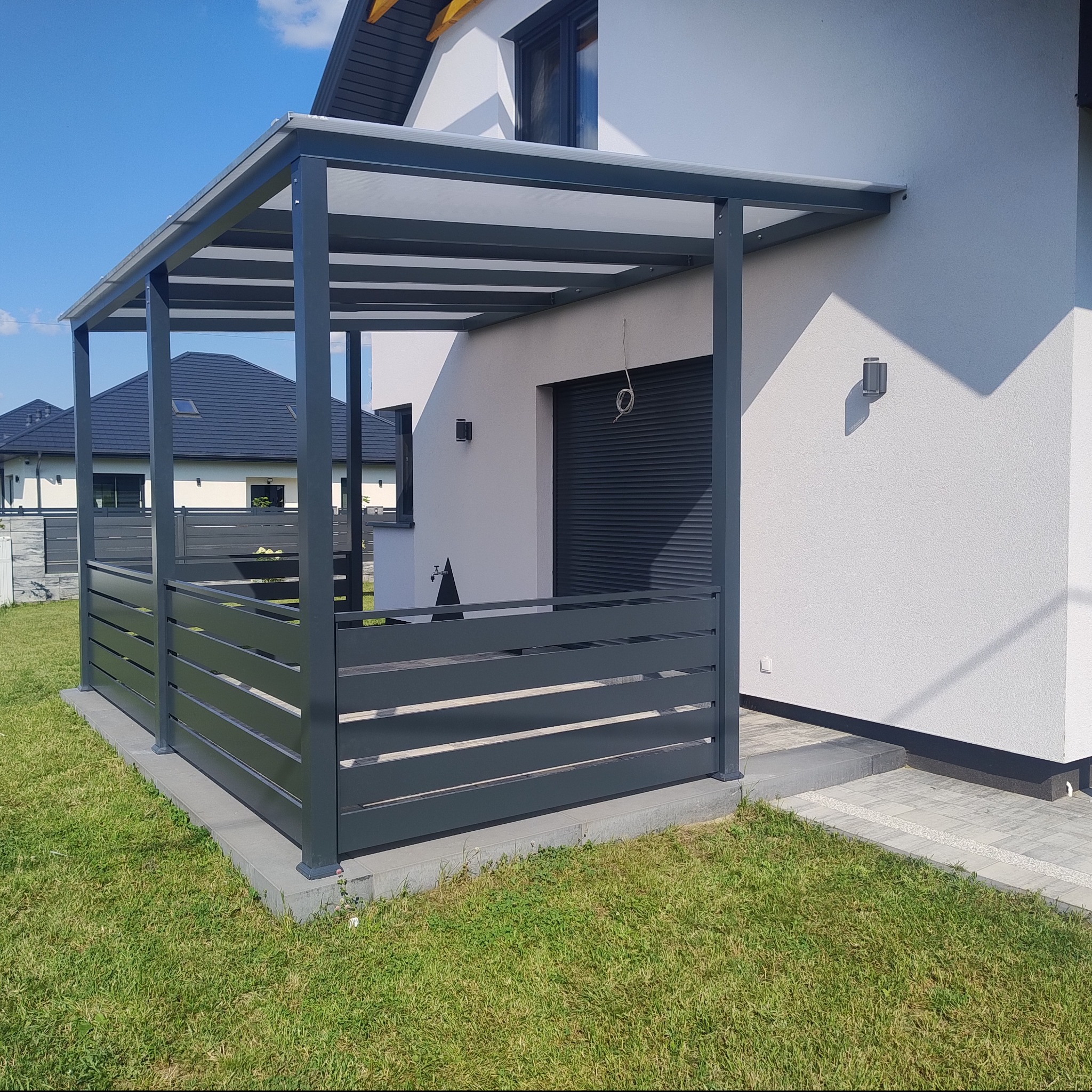 Nowoczesna, szara konstrukcja zadaszenia tarasu z poziomymi panelami w ogrodzie, przylegająca do białego domu z roletami zewnętrznymi.