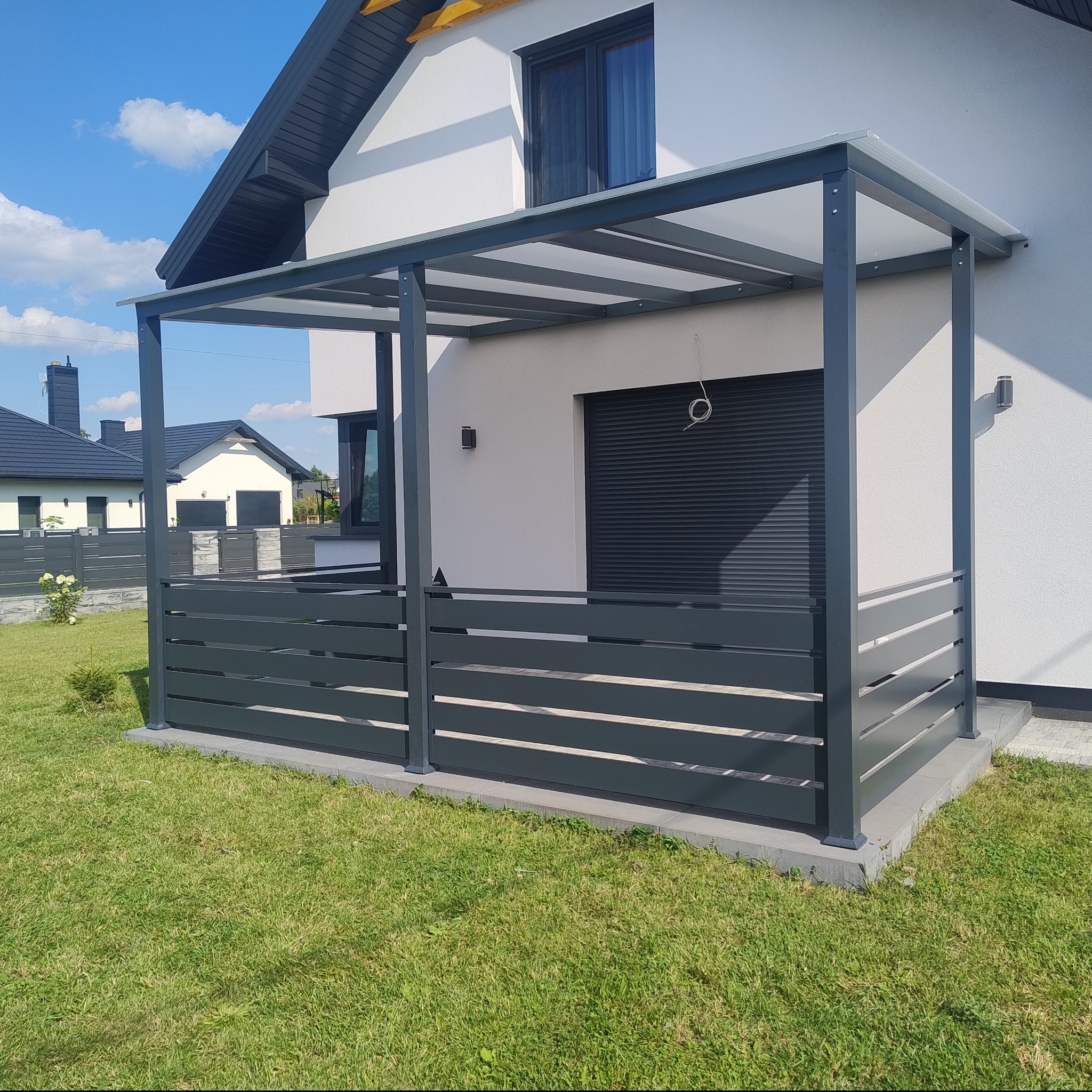 Nowoczesna, antracytowa pergola z poziomymi panelami na tarasie przylegającym do białego domu z szarą stolarką okienną i roletami zewnętrznymi, na tle zielonego trawnika i sąsiednich domów...