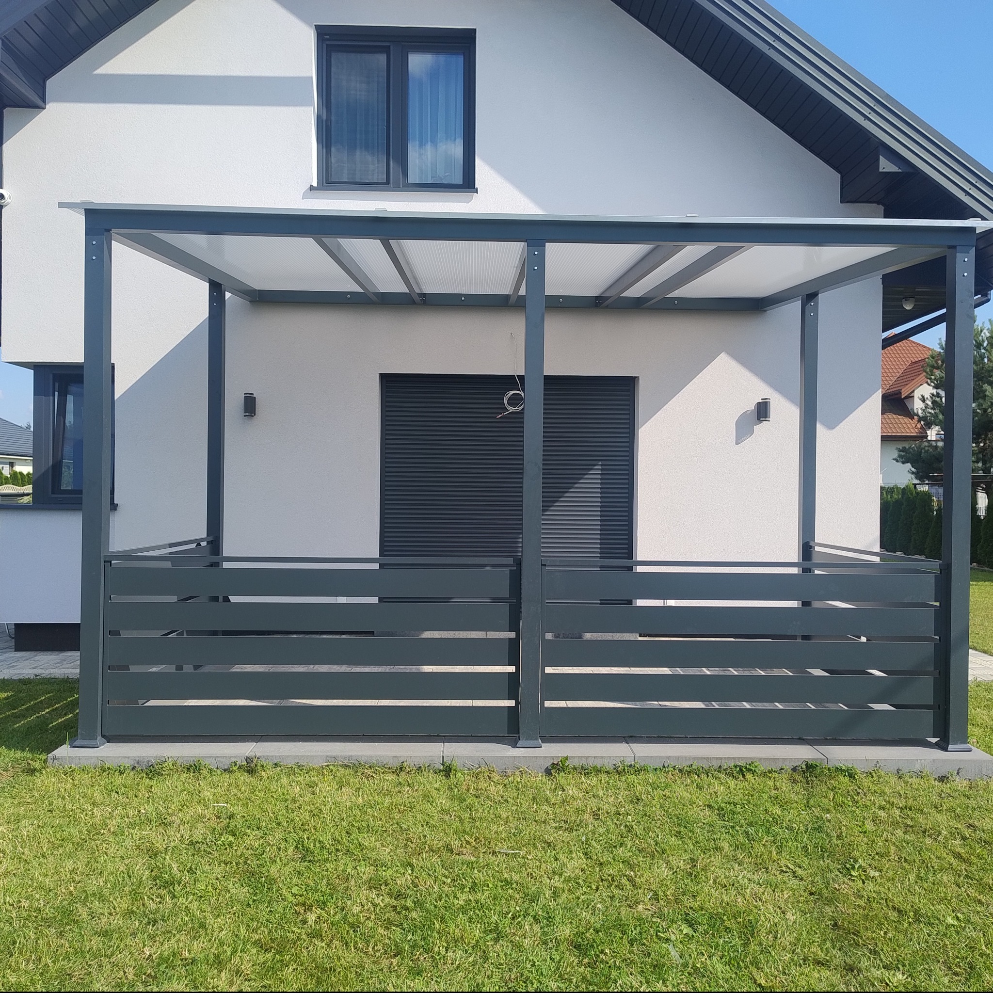 Nowoczesna, szara pergola z metalową balustradą i przezroczystym zadaszeniem na tle białej ściany domu z zamkniętą żaluzją.