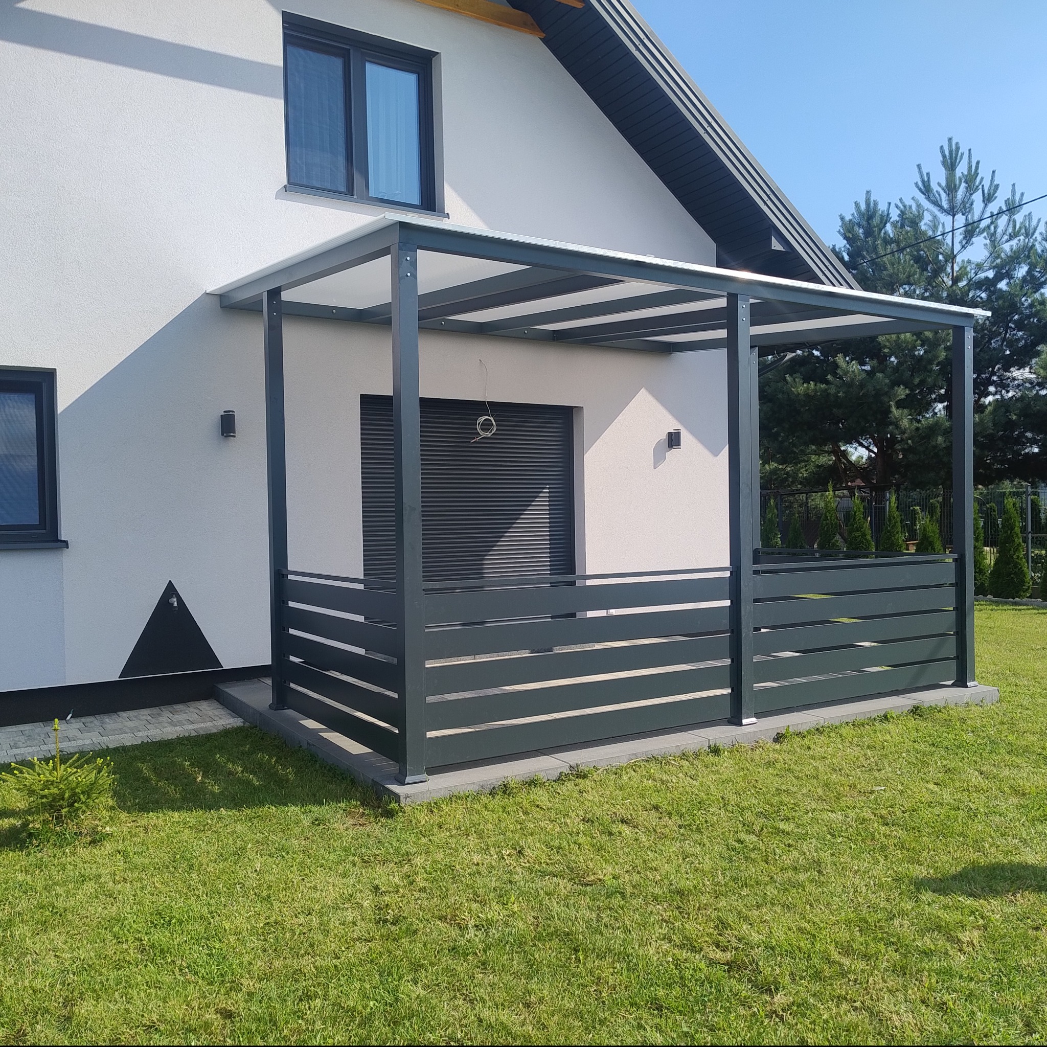 Nowoczesna, szara konstrukcja z metalu, pełniąca funkcję zadaszonego tarasu, przylegająca do białego domu z szarą stolarką okienną i roletami zewnętrznymi, otoczona zielonym trawnikiem i drzewami.