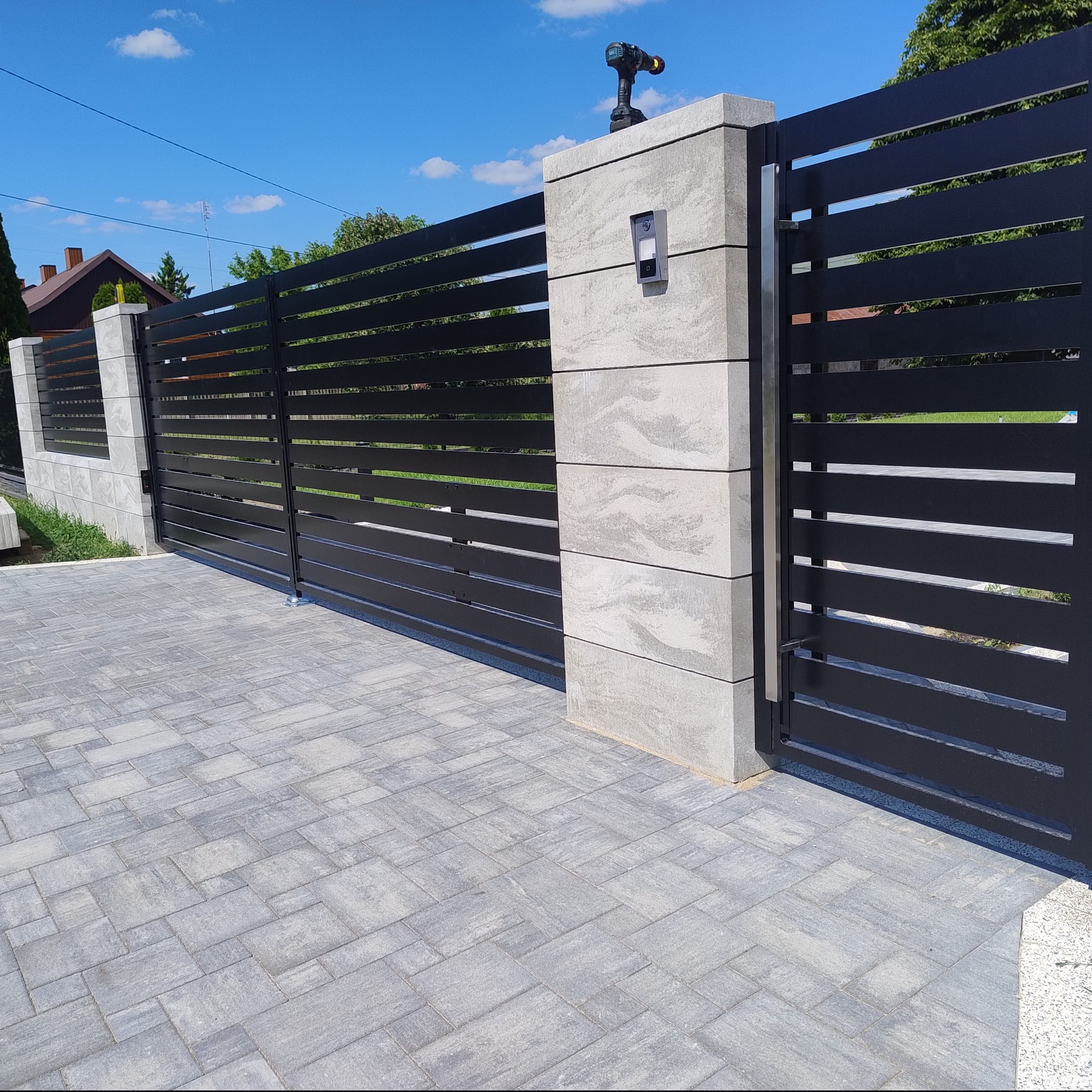 Nowoczesna, pozioma, czarna brama wjazdowa z jasnoszarymi, betonowymi słupkami, na jednym ze słupków zamontowany jest domofon, a na jego szczycie leży wkrętarka akumulatorowa.