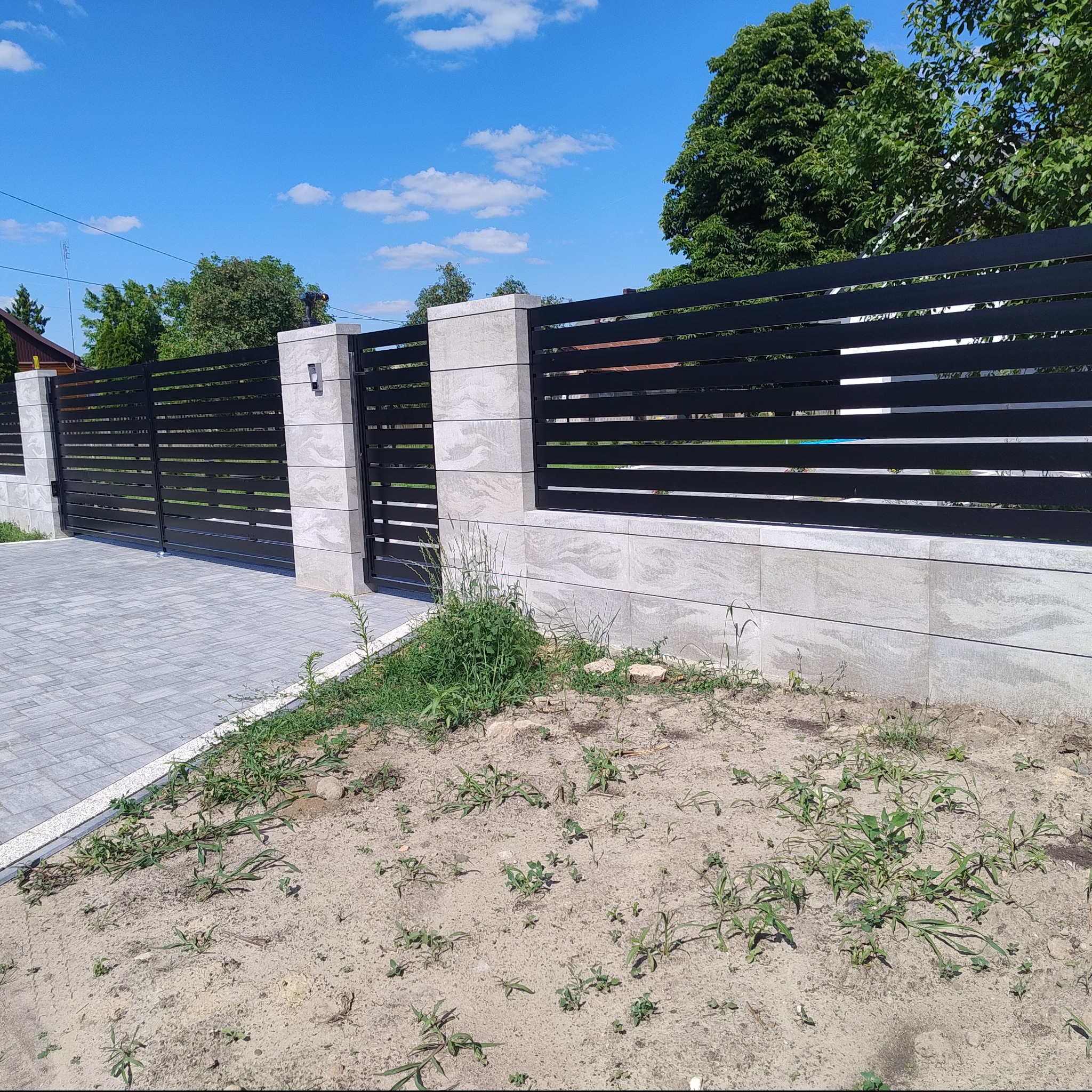 Nowoczesne ogrodzenie posesji z poziomych, czarnych profili metalowych na niskiej podmurówce z szarej kostki, zakończone słupkami z oświetleniem, widok na niezagospodarowany teren przed ogrodzeniem.