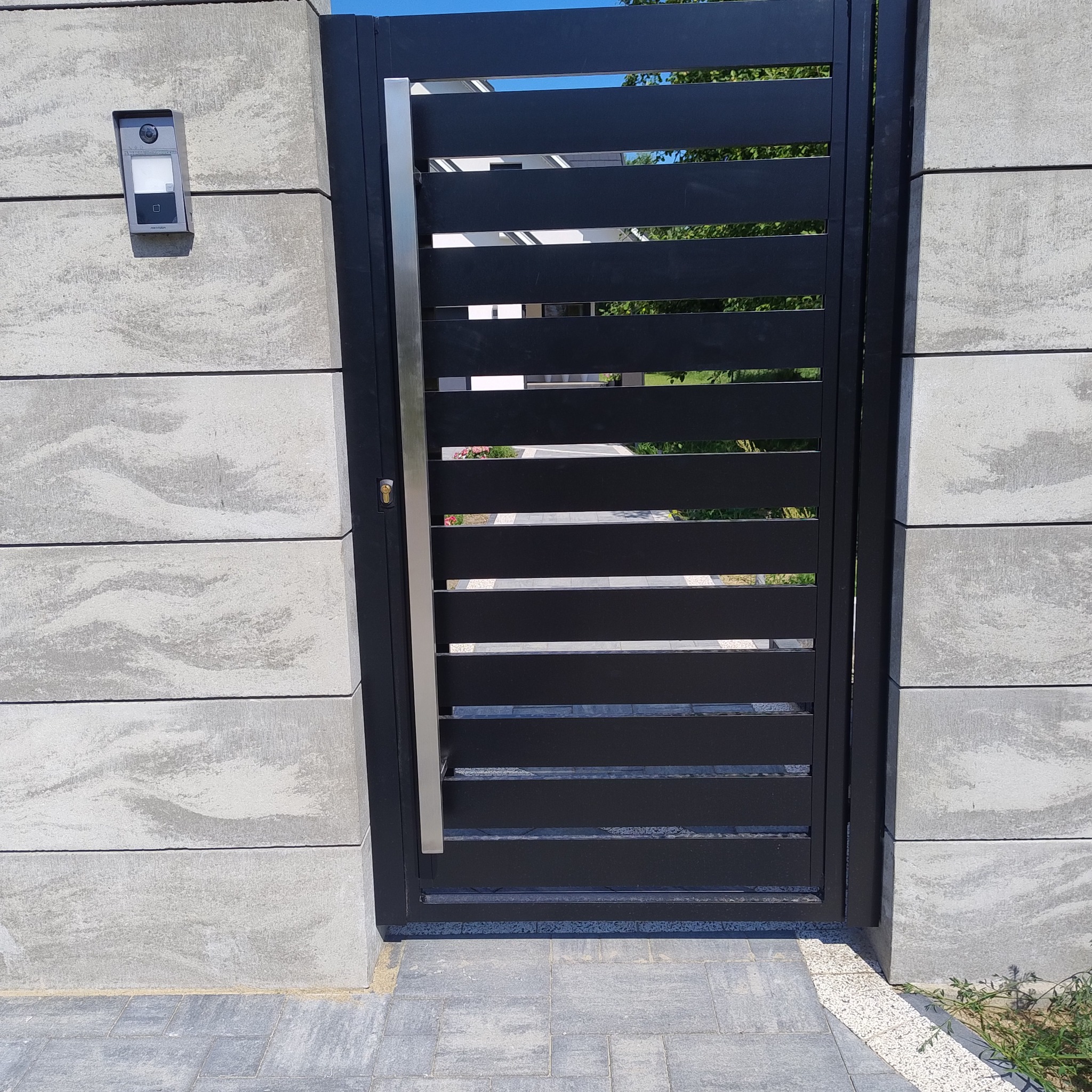 Nowoczesna, czarna furtka z poziomymi szczeblami i pionowym, srebrnym uchwytem, zamontowana w szarym, betonowym ogrodzeniu z wideodomofonem po lewej stronie. Widok na posesję przez prześwity między...