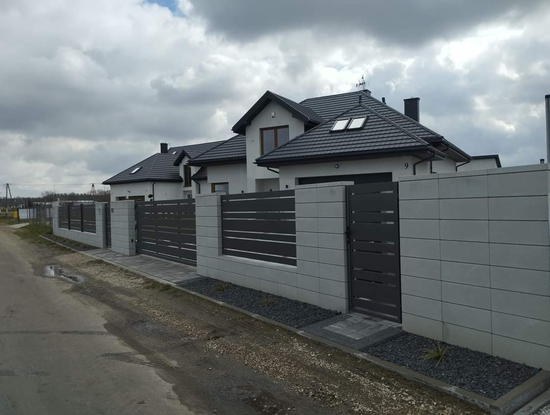 Nowoczesne ogrodzenie posesji z szarego betonu i poziomych, metalowych paneli w kolorze grafitowym, dopasowane do architektury domu z ciemną dachówką, widok z ulicy w pochmurny dzień.