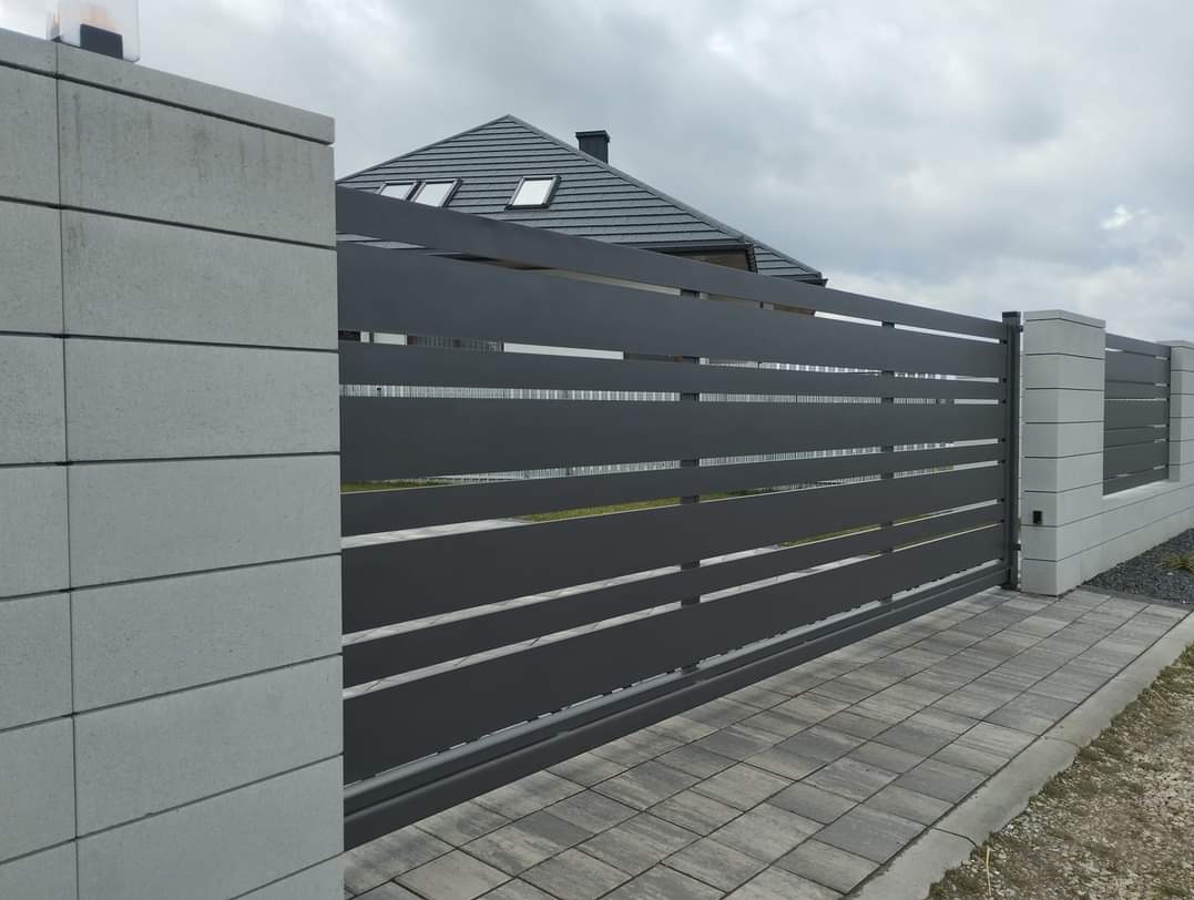 Nowoczesna, przesuwna brama w kolorze antracytowym z poziomymi szczelinami, zamontowana na betonowych słupkach wykończonych szarym kamieniem, na tle domu z szarą dachówką i pochmurnego nieba.