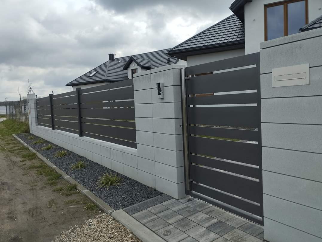 Nowoczesna brama wjazdowa i ogrodzenie metalowe z poziomymi panelami, osadzone na betonowych słupkach i podmurówce, z ozdobnym kamiennym obrzeżem rabaty.