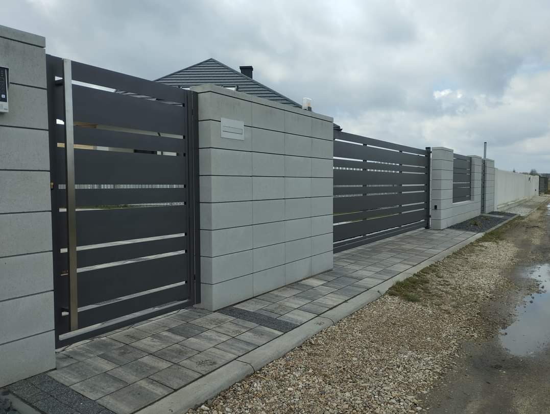 Nowoczesna, przesuwna brama wjazdowa z poziomymi panelami w kolorze grafitowym, osadzona w betonowych słupkach z szarej kostki, widok z zewnątrz posesji, obok chodnik z kostki brukowej.