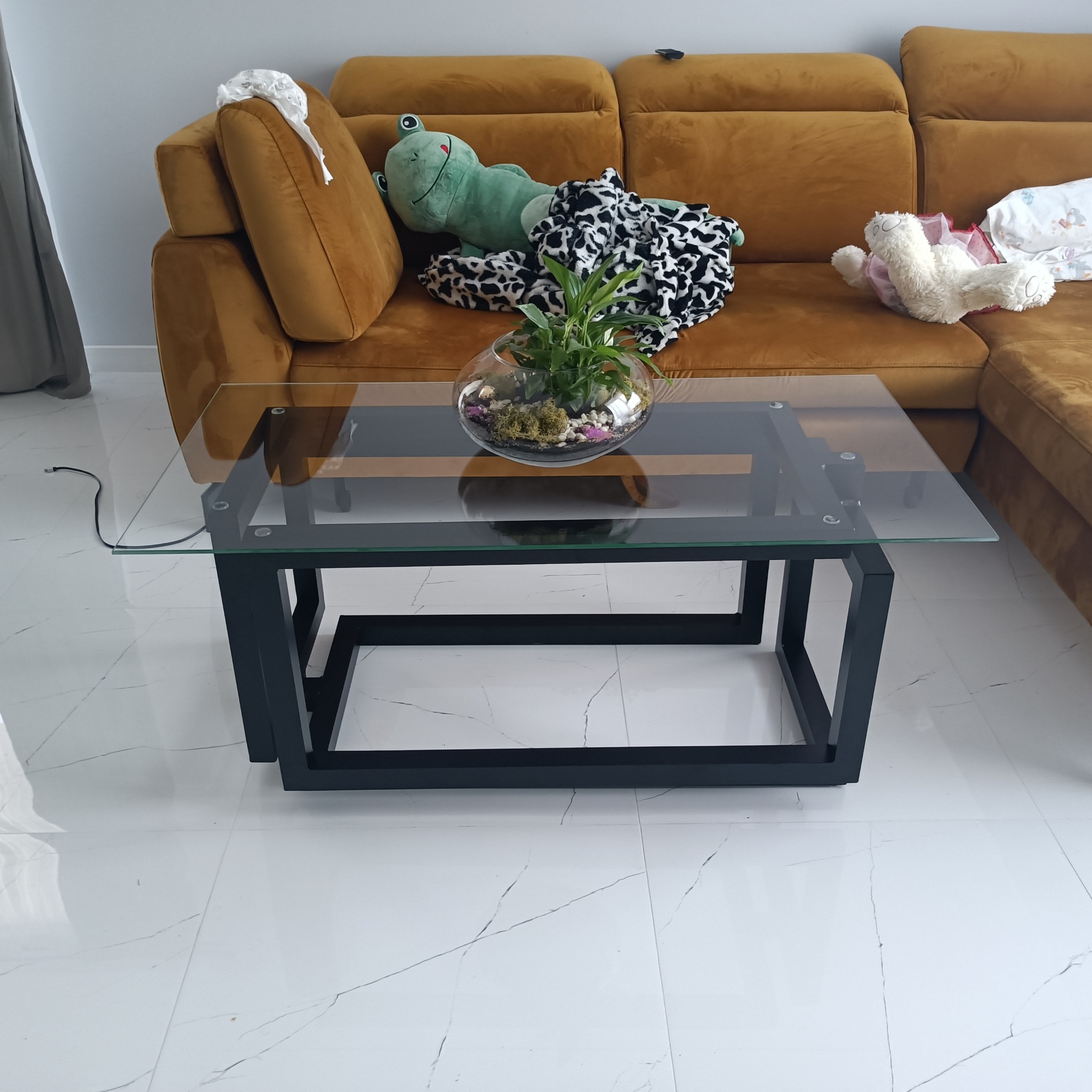 Stolik kawowy z czarną metalową podstawą i szklanym blatem, na którym stoi szklana kula z roślinami, w tle musztardowa sofa z pluszakami i kocem.