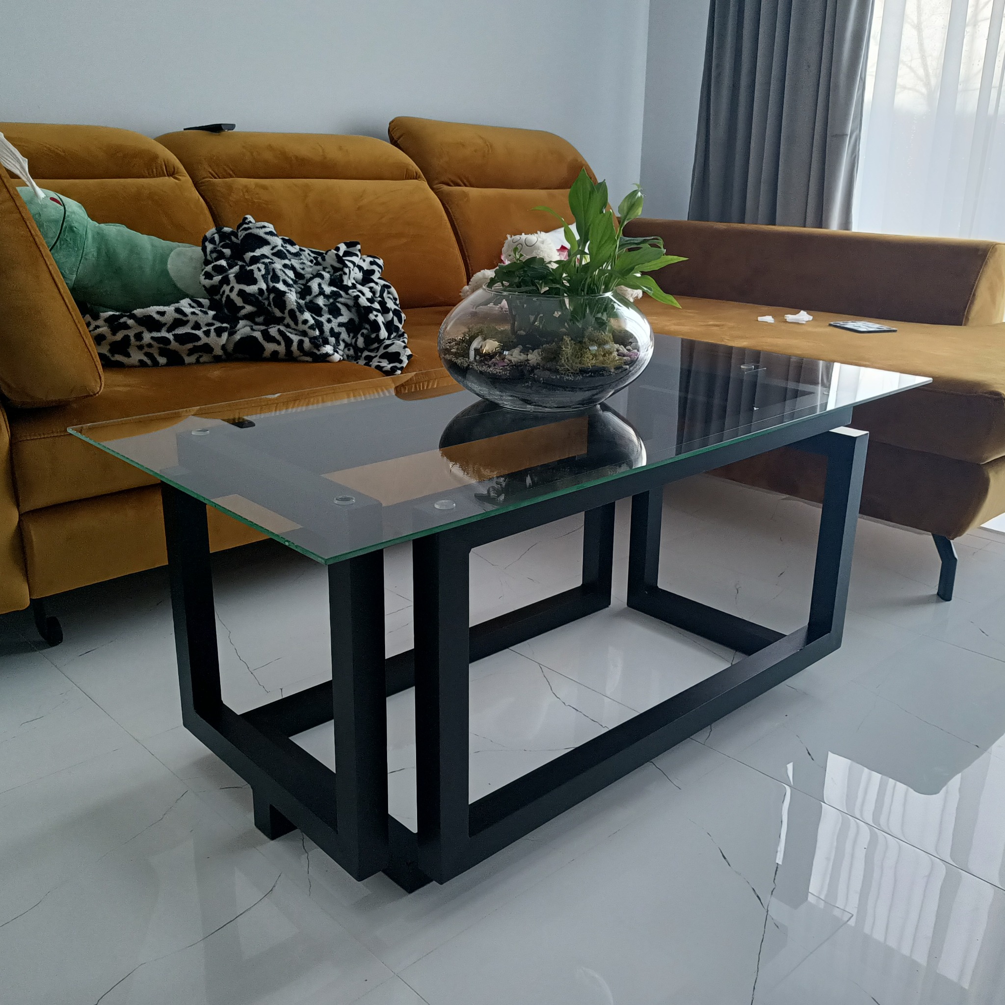 Stolik kawowy z czarną, metalową podstawą i szklanym blatem, na którym stoi szklana kula z roślinami, na tle musztardowej sofy i białych zasłon.