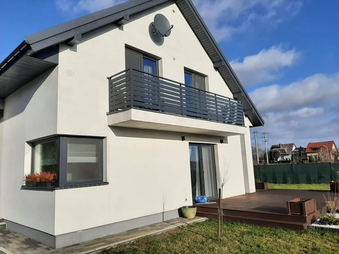 Nowoczesny dom z balkonem z szarą metalową balustradą poziomą, widok z ogrodu, drewniany taras i zadbany trawnik.