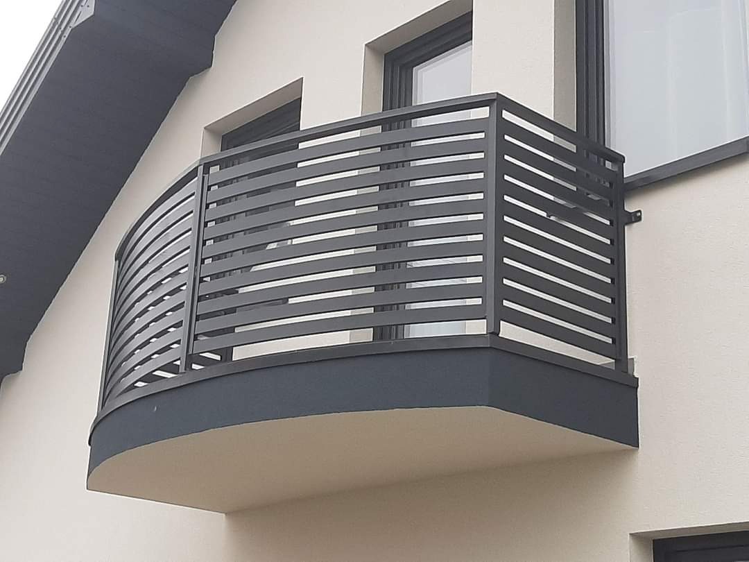 Szara, metalowa balustrada balkonowa z poziomymi szczeblami, zamontowana na zaokrąglonym balkonie nowoczesnego domu.