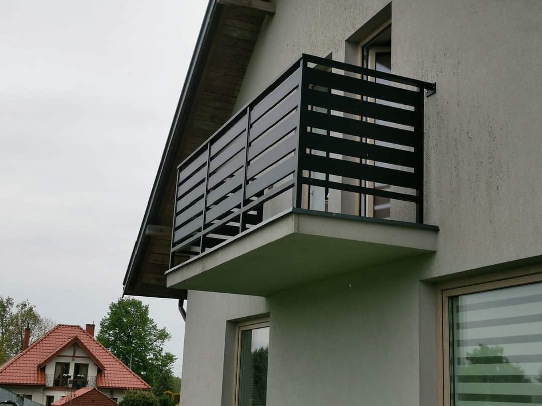 Nowoczesny balkon z czarną, poziomą balustradą z metalowych profili, widok z dołu, w tle dom z czerwoną dachówką.