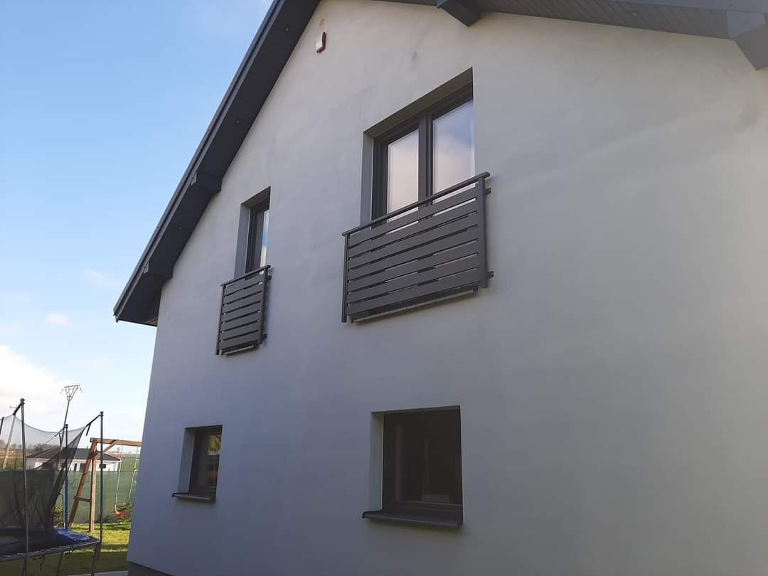 Elewacja domu z dwoma balkonami francuskimi zabezpieczonymi szarymi, poziomymi barierkami. W tle widoczny plac zabaw.