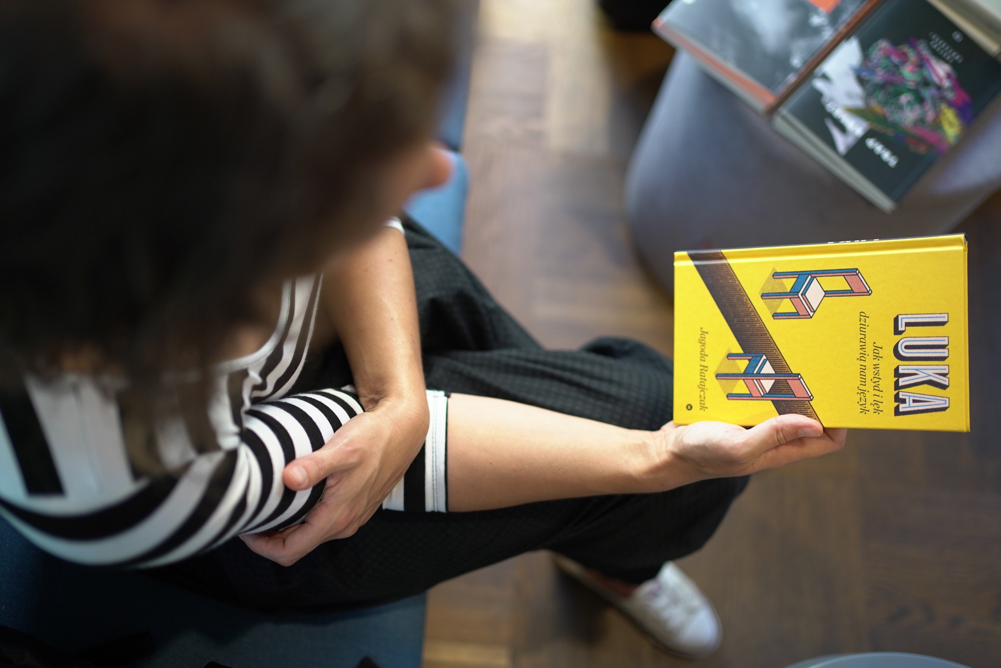 Osoba w bluzie w paski trzyma żółtą książkę 'LUKA' Jagody Ratajczak z ilustracjami geometrycznych krzeseł na okładce, siedząc na niebieskim krześle, obok stosu innych książek.