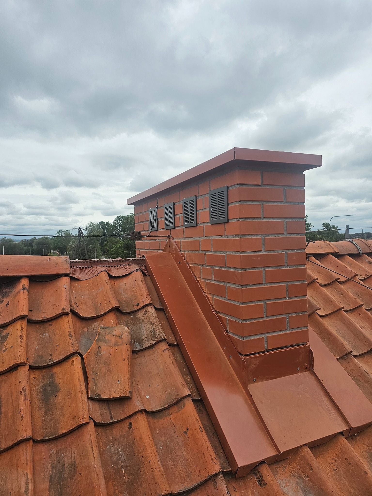 Ceglany komin z metalową obróbką na dachu z dachówki ceramicznej, widoczne kratki wentylacyjne, pochmurne niebo w tle.