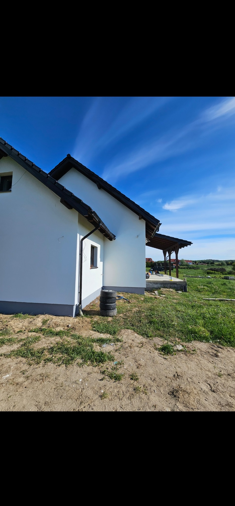 Świeżo otynkowany dom jednorodzinny z czarnym dachem i stosem opon przy ścianie, widok na drewnianą altanę w tle.
