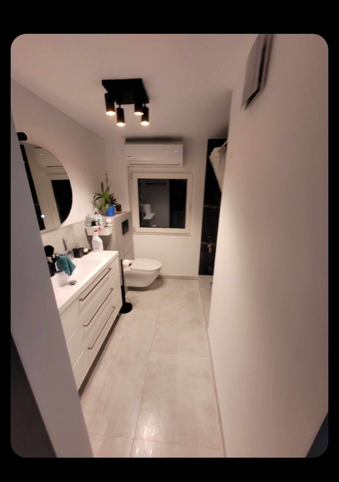 Nowoczesna łazienka z białymi meblami, okrągłym lustrem, podwieszaną toaletą i oświetleniem punktowym na suficie.