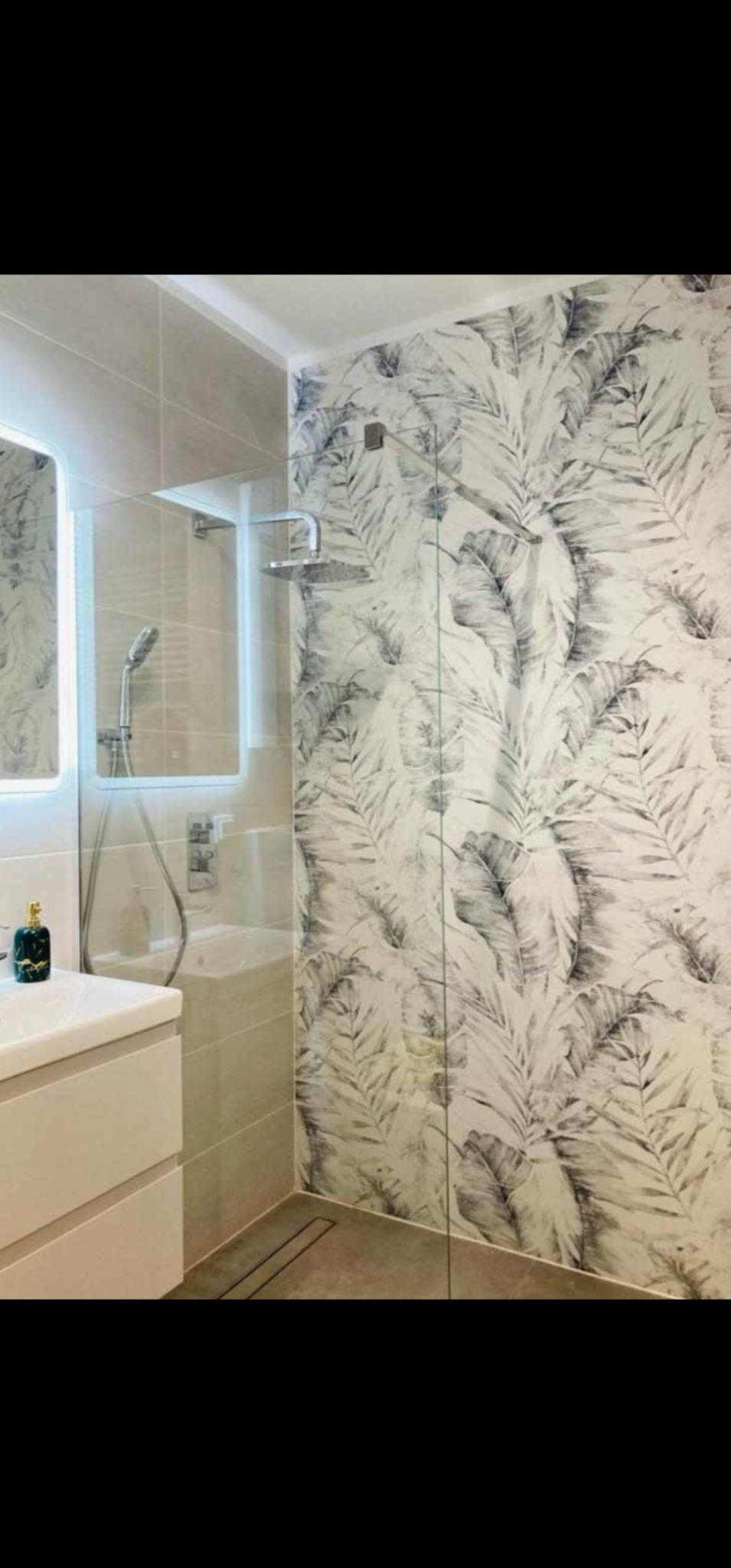 Nowoczesna łazienka z przeszkloną kabiną prysznicową, jasnymi płytkami imitującymi kamień, ścianą akcentową z tapetą w motywie liści palmowych oraz podświetlanym lustrem.