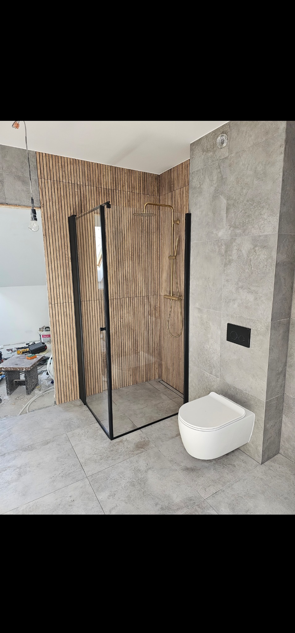 Nowoczesna łazienka w trakcie wykończenia: kabina prysznicowa z czarnymi profilami i złotą armaturą na tle drewnianej ściany z lamel, podwieszana toaleta na szarej ścianie z płytek, widoczne...