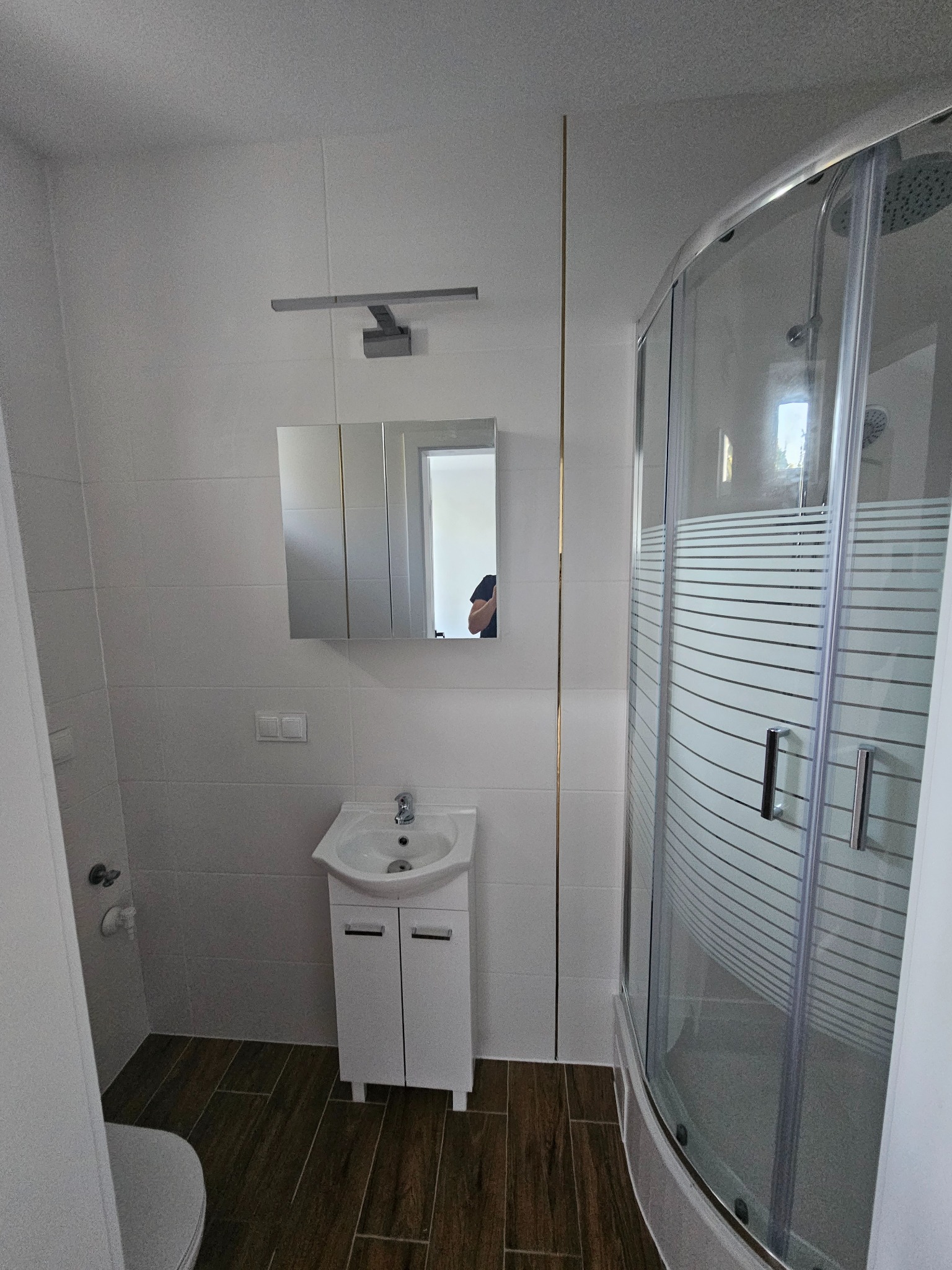 Nowoczesna łazienka z kabiną prysznicową z poziomymi pasami, białą umywalką z szafką, lustrem z oświetleniem i brązowymi płytkami podłogowymi.