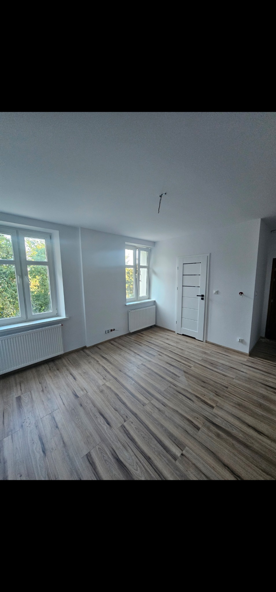 Jasne panele podłogowe ułożone w pustym pokoju z białymi ścianami, grzejnikami pod oknami i białą, nowoczesną stolarką drzwiową.