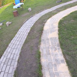 Clean House Jaworski Przemysław - Ścieżka z kostki brukowej wijąca się przez trawnik. Część ścieżki jest już ułożona, a część przygotowana pod układanie. Widoczne narzędzia i materiały budowlane w tle.