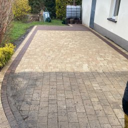 Clean House Jaworski Przemysław - Ułożona kostka brukowa na podjeździe, z widocznym fragmentem czyszczonej powierzchni. W tle budynek z białymi ścianami i zielona roślinność.