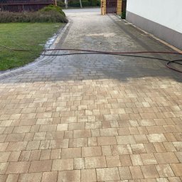 Clean House Jaworski Przemysław - Częściowo umyta kostka brukowa na podjeździe, widoczna różnica w kolorze. W tle trawnik, budynek i brama wjazdowa. Wąż ciśnieniowy na kostce.