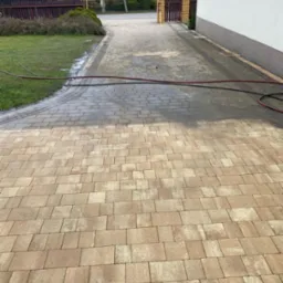Częściowo umyta kostka brukowa na podjeździe, widoczna różnica w kolorze. W tle trawnik, budynek i brama wjazdowa. Wąż ciśnieniowy na kostce.